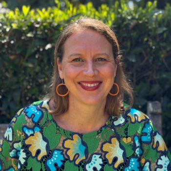 Outdoor headshot of a smiling woman wearing orange earrings and a colourful patterned top, with greenery in the background.